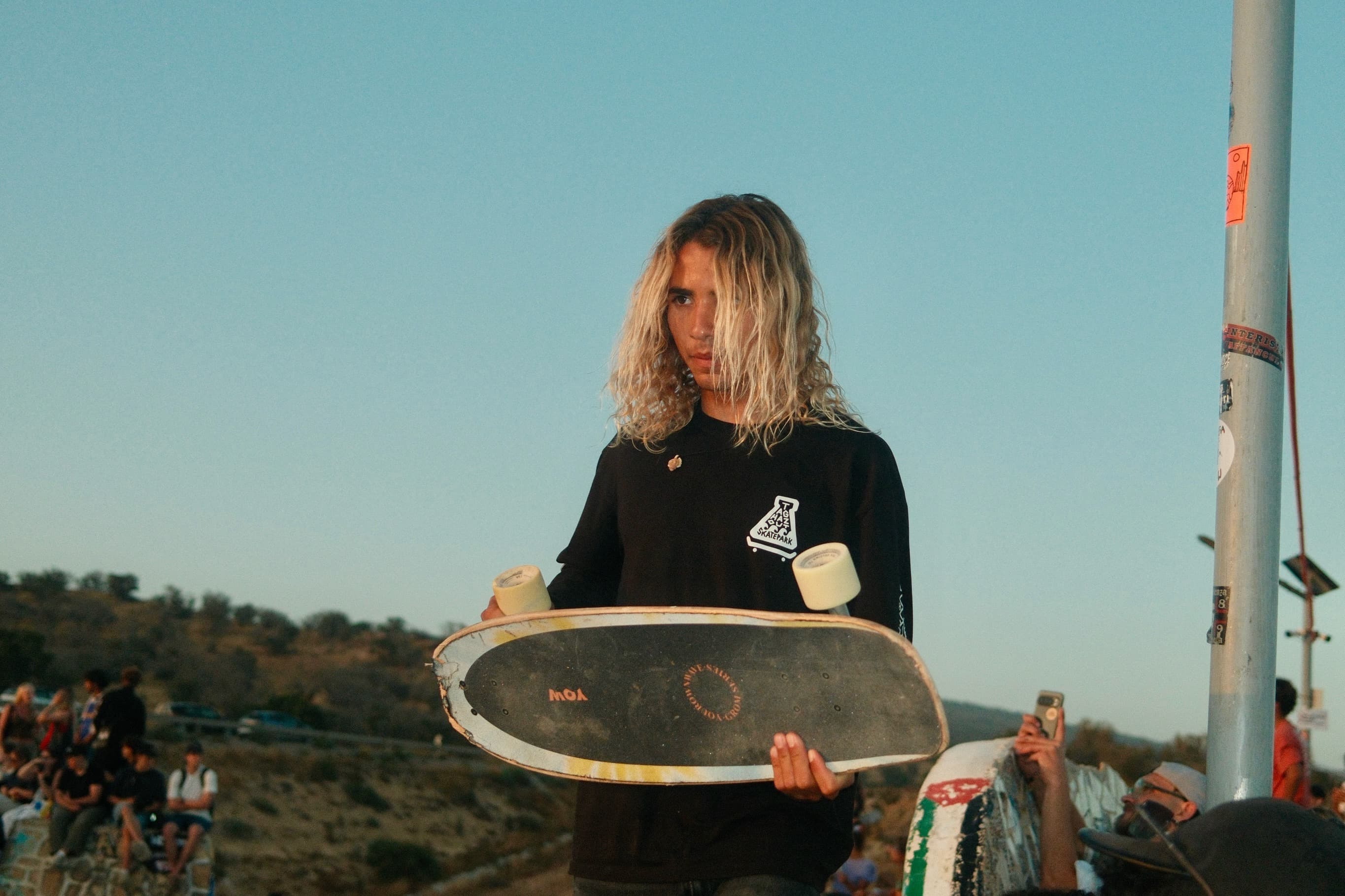 Skater holding a worn deck at the park in the black Taghazout Skatepark long sleeve, chest patch and sleeve print in view