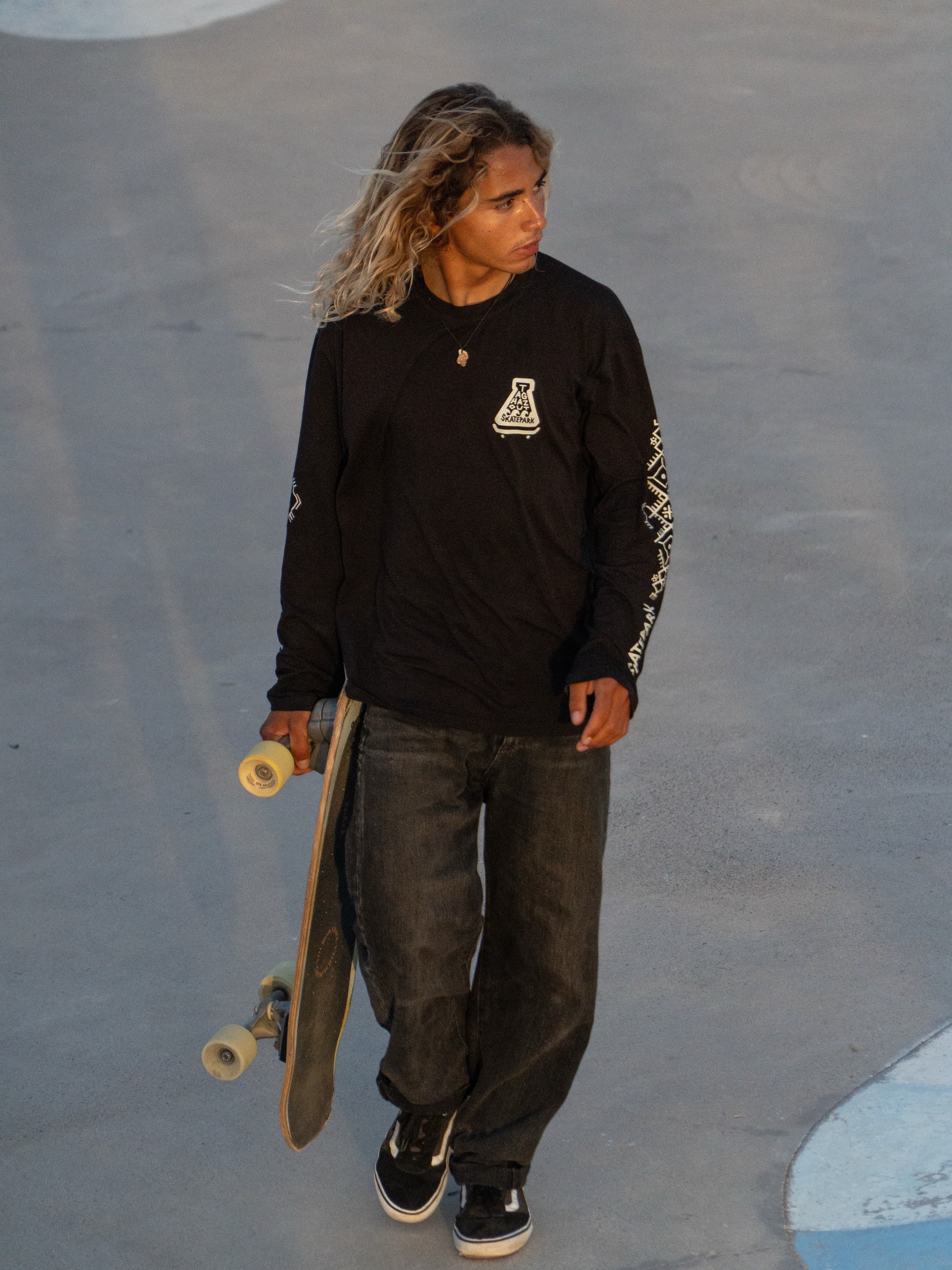Skater in the black Taghazout Skatepark long sleeve, board in hand, crossing the park at golden hour