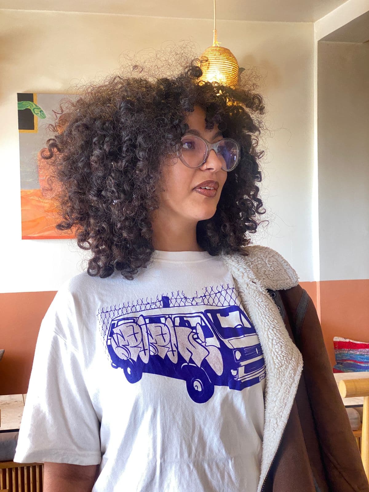 Wearing the white van-print tee under a shearling jacket, indoors at the studio